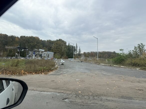 IMG_8881 Desolate roadside with scattered debris and a ruined metal structure by an overgrown lot under a cloudy sky.