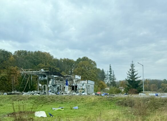 IMG_8882 Ruined metal-framed structure with scattered debris in a grassy field under a cloudy sky, surrounded by trees in the distance.