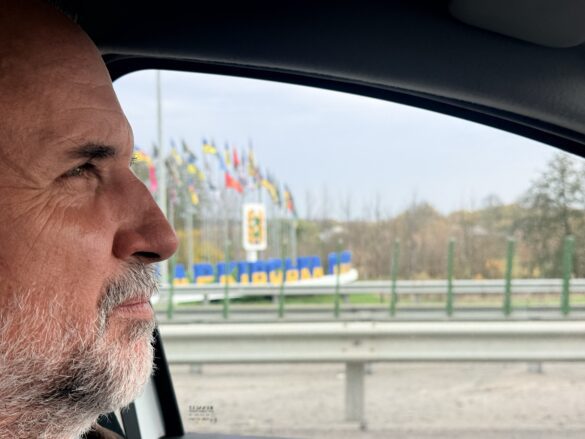 IMG_8885 Side profile of an older man with a gray beard sitting in a car, looking out the window at blurred flags and a guardrail on the highway.