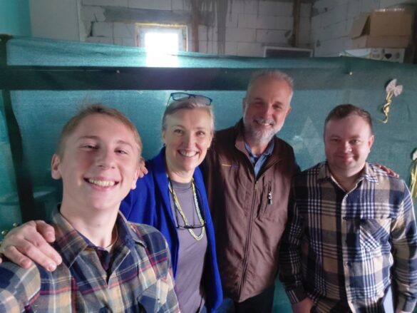 IMG_8920 Four adults posing for a group selfie in a workshop, smiling at the camera, wearing casual plaid shirts and jackets over their outfits.