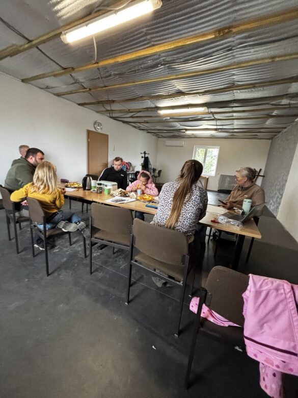 IMG_8981 People seated around a long table in a unfinished room, sharing a meal and chatting. Loose construction ceiling and bright overhead lights visible in the background.