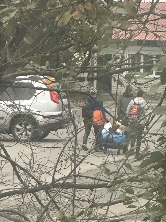 IMG_9351 People on a residential street beside a white car, seen through tree branches; one person wears an orange backpack.