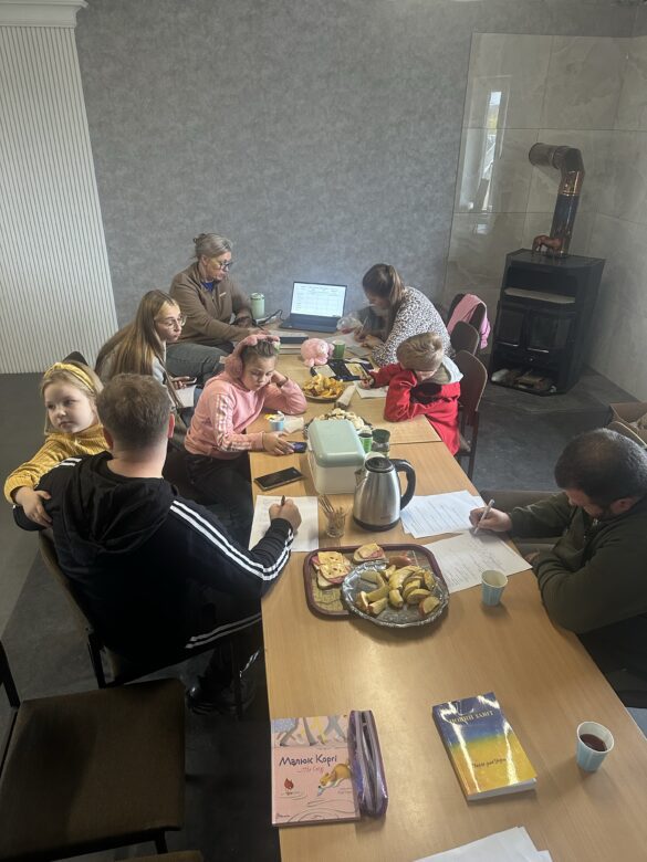 IMG_9354 Group of adults and children seated around a long table in a cozy room, working on papers with snacks and a laptop.