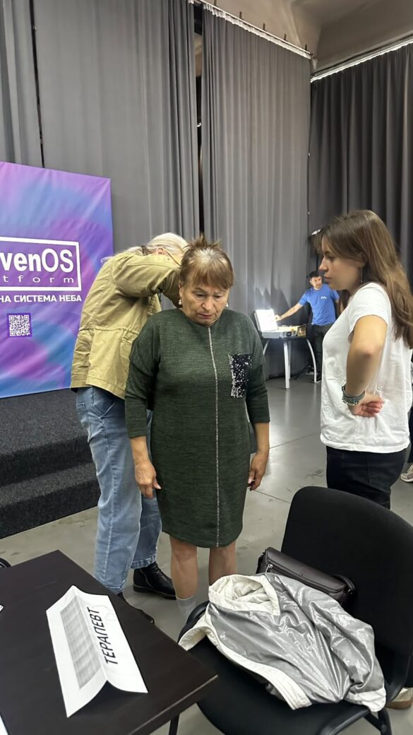 Older woman in a dark green dress stands with hands at her sides, while a younger woman in a white shirt speaks to her backstage next to gray curtains and a purple event banner.