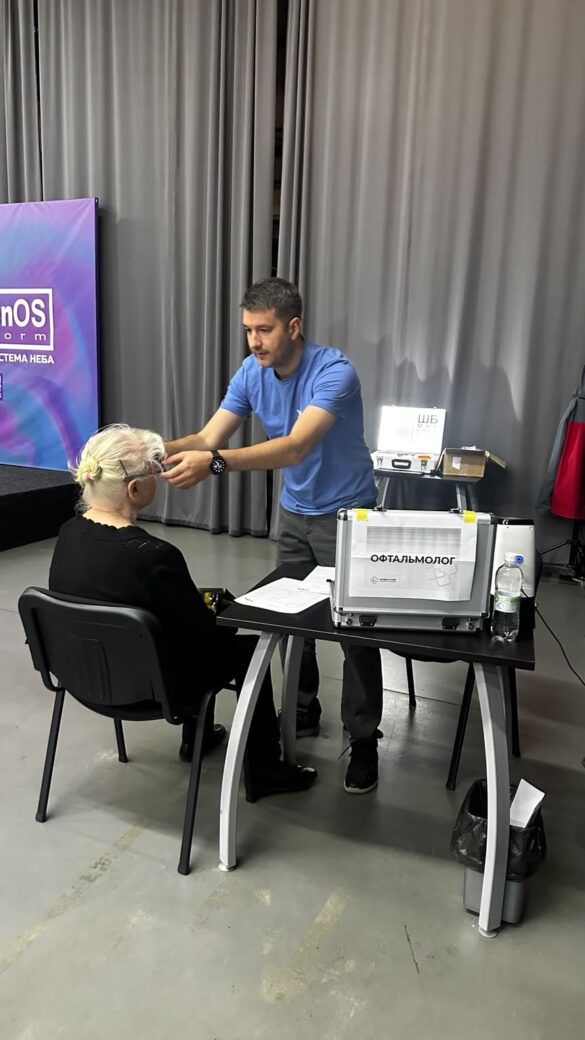 Clinician in a blue shirt conducts an eye exam for an elderly patient seated at a table with ophthalmology equipment nearby.
