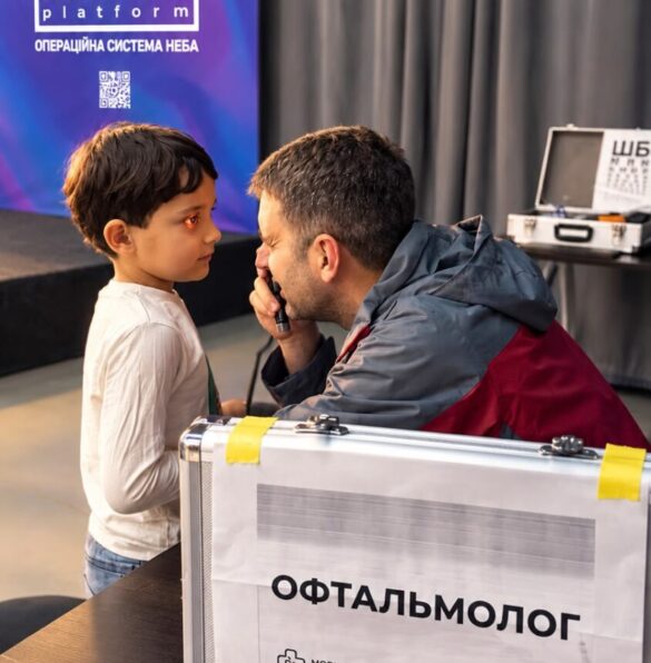 Child speaking with an ophthalmologist during a medical screening at an event; a labeled ophthalmology case sits nearby.