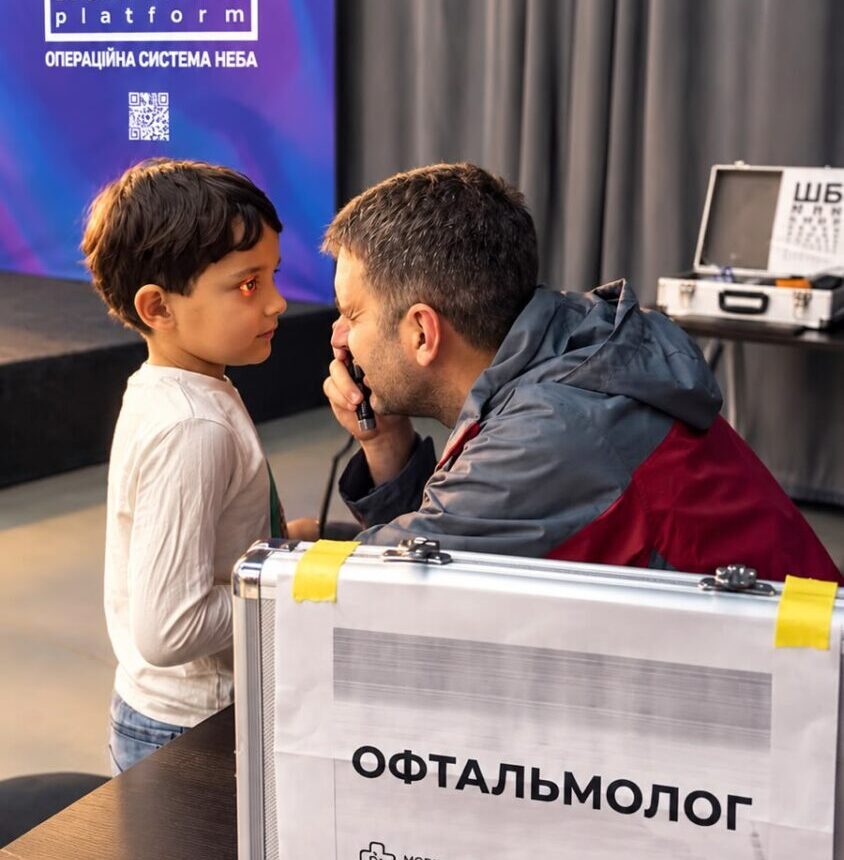 Child speaking with an ophthalmologist during a medical screening at an event; a labeled ophthalmology case sits nearby.
